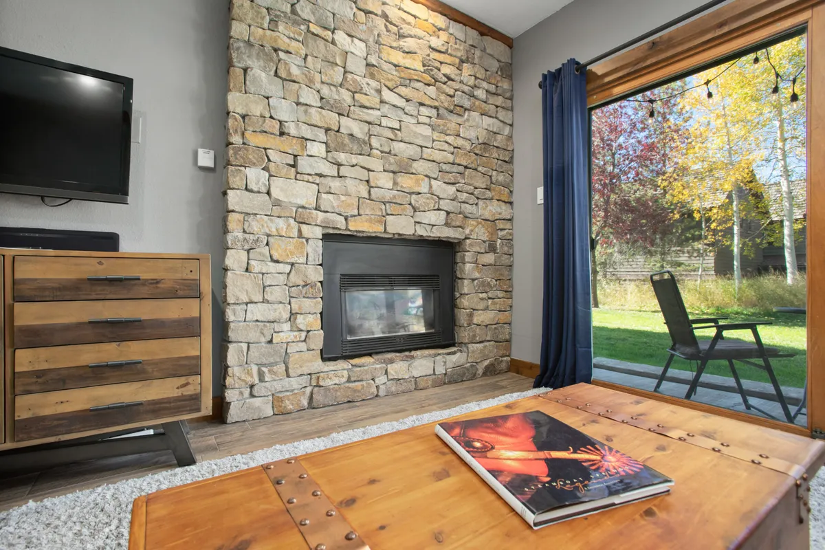 This image presents a warm and inviting living room with a stone fireplace as the focal point, surrounded by plush seating and wooden furniture. Large glass doors lead directly to the outdoors, and a coffee table with a magazine adds a lived-in feel. The soft natural light coming through emphasizes the open, airy layout and the close connection to the patio and yard.