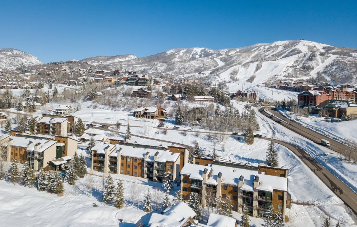A stunning mountain landscape featuring snow-capped peaks, modern buildings nestled among pine trees, and a winding road leading into adventure. The scene evokes the promise of outdoor activities like skiing, snowshoeing, or hiking right from the doorstep.