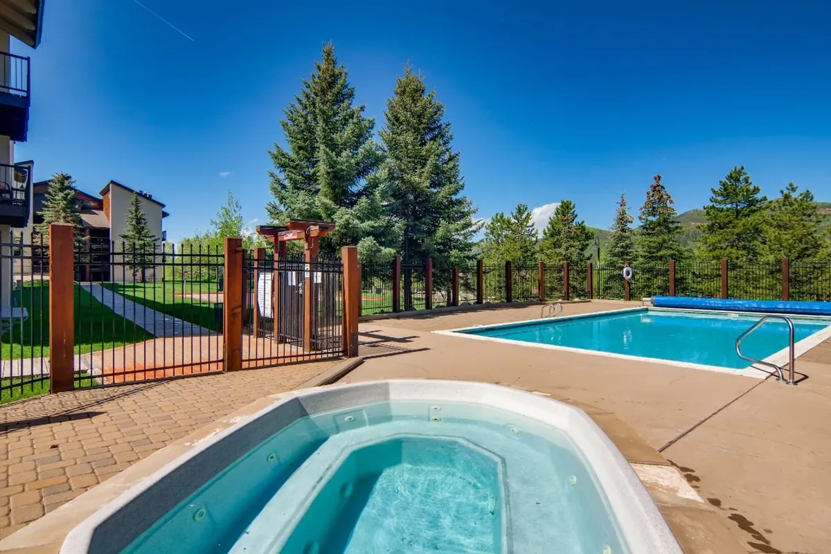 A tranquil outdoor scene showcasing a jacuzzi and swimming pool surrounded by trees. The area is fenced for privacy and offers both sun and shaded spots for relaxation. The water sparkles under natural light, and the space is set up for both intimate relaxation and social gatherings.