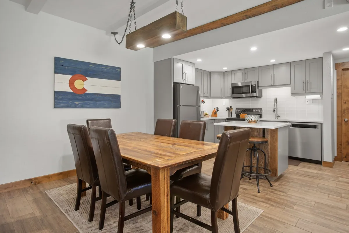 The image displays the open-plan dining and kitchen area, highlighting the all-on-one-level living arrangement. It features a dining table, kitchen appliances, and Colorado flag artwork, creating an inviting and functional space for both short and long stays.