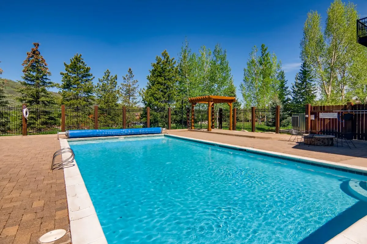 A beautiful exterior scene featuring a sparkling swimming pool surrounded by lush greenery, a pergola, and a cozy fire pit area. The poolside space looks inviting and is set up for social gatherings, relaxation, and family fun. The setting suggests warm, lively summer days and evenings spent outdoors.