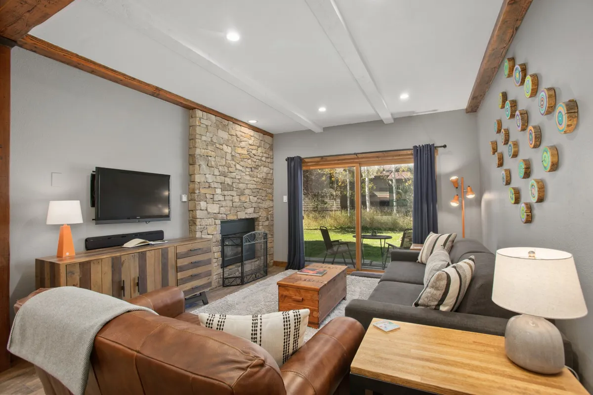 A stylish and inviting condo living room featuring a stone fireplace, comfortable lounge seating, tasteful artistic wall decor, and expansive windows that frame serene nature views. The space is bathed in natural light, creating a warm and welcoming atmosphere perfect for relaxing after a day on the slopes.