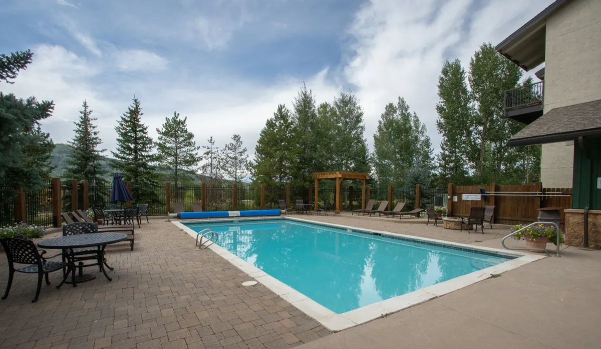 A tranquil outdoor pool area surrounded by trees, a pergola, and comfortable seating. The space feels social yet serene, ideal for relaxation, sunbathing, or gathering with neighbors and friends.