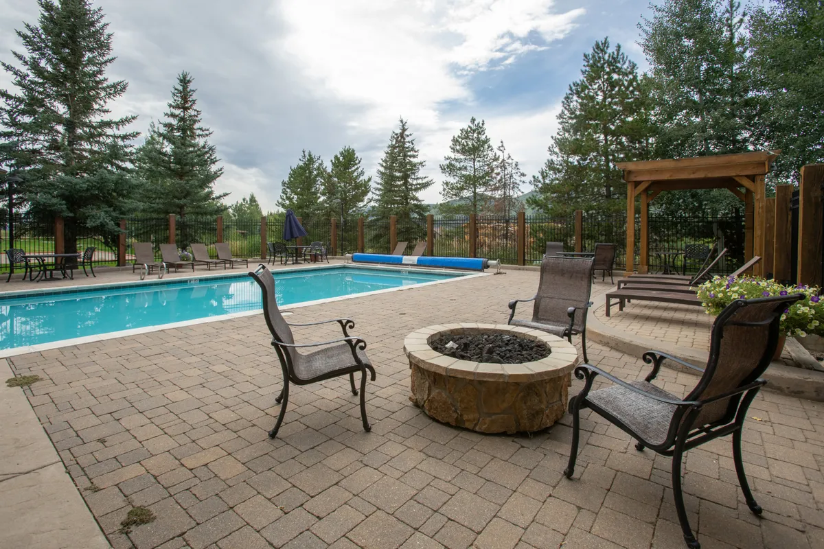 The photo highlights a sunlit pool area surrounded by trees, featuring a fire pit, deck chairs, and ample space for group gatherings. The setting evokes social summer evenings, with possibilities for swimming, lounging, and enjoying warm nights around the fire pit, all within a lush green environment.