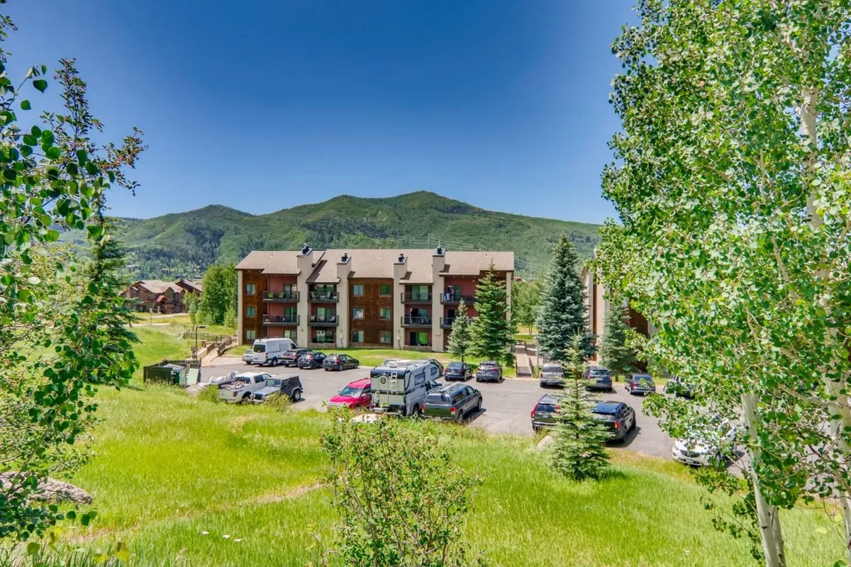This image showcases the main building of The Rockies complex, surrounded by lush green trees and set against a stunning mountain backdrop. The parking area and the natural environment highlight accessibility and a scenic setting, inviting guests to explore the neighborhood and enjoy the outdoors.