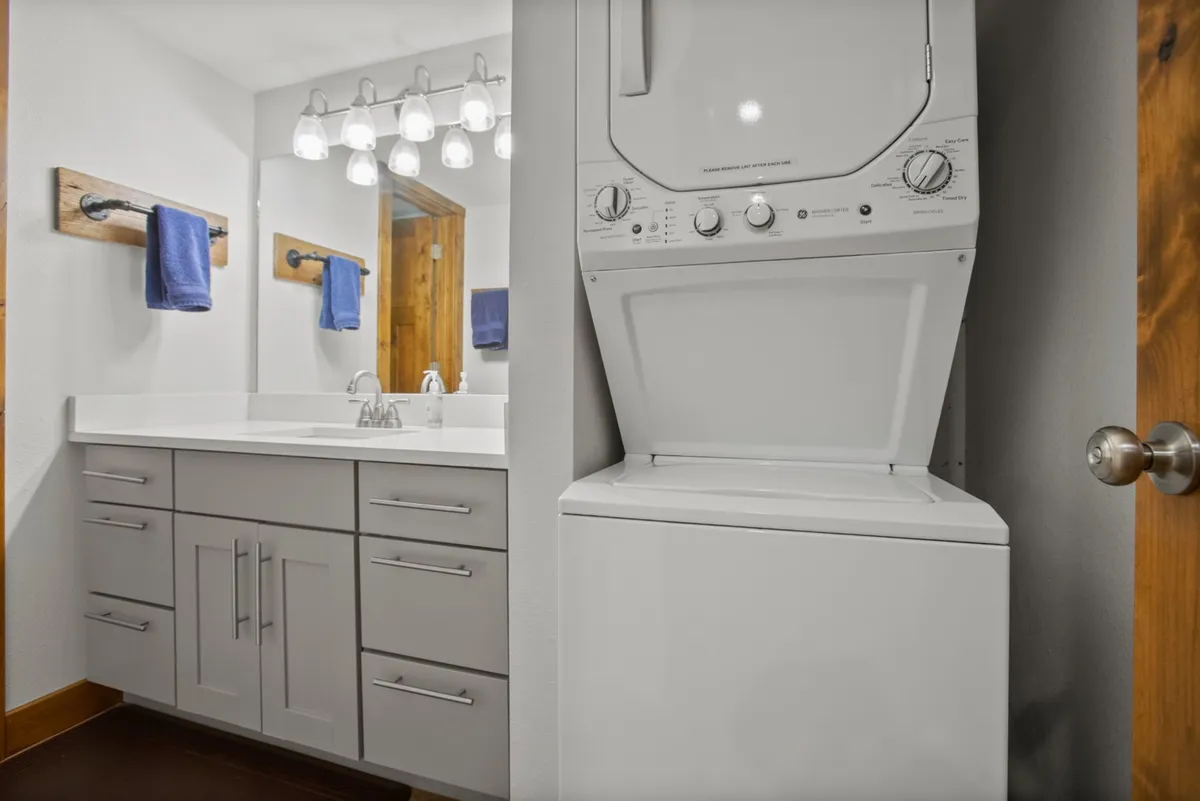A spacious bathroom with a modern vanity and illuminated mirror, complemented by a stackable washer and dryer unit. Fresh hand towels and a clean, organized setup emphasize functionality and comfort, supporting multitasking for guests.