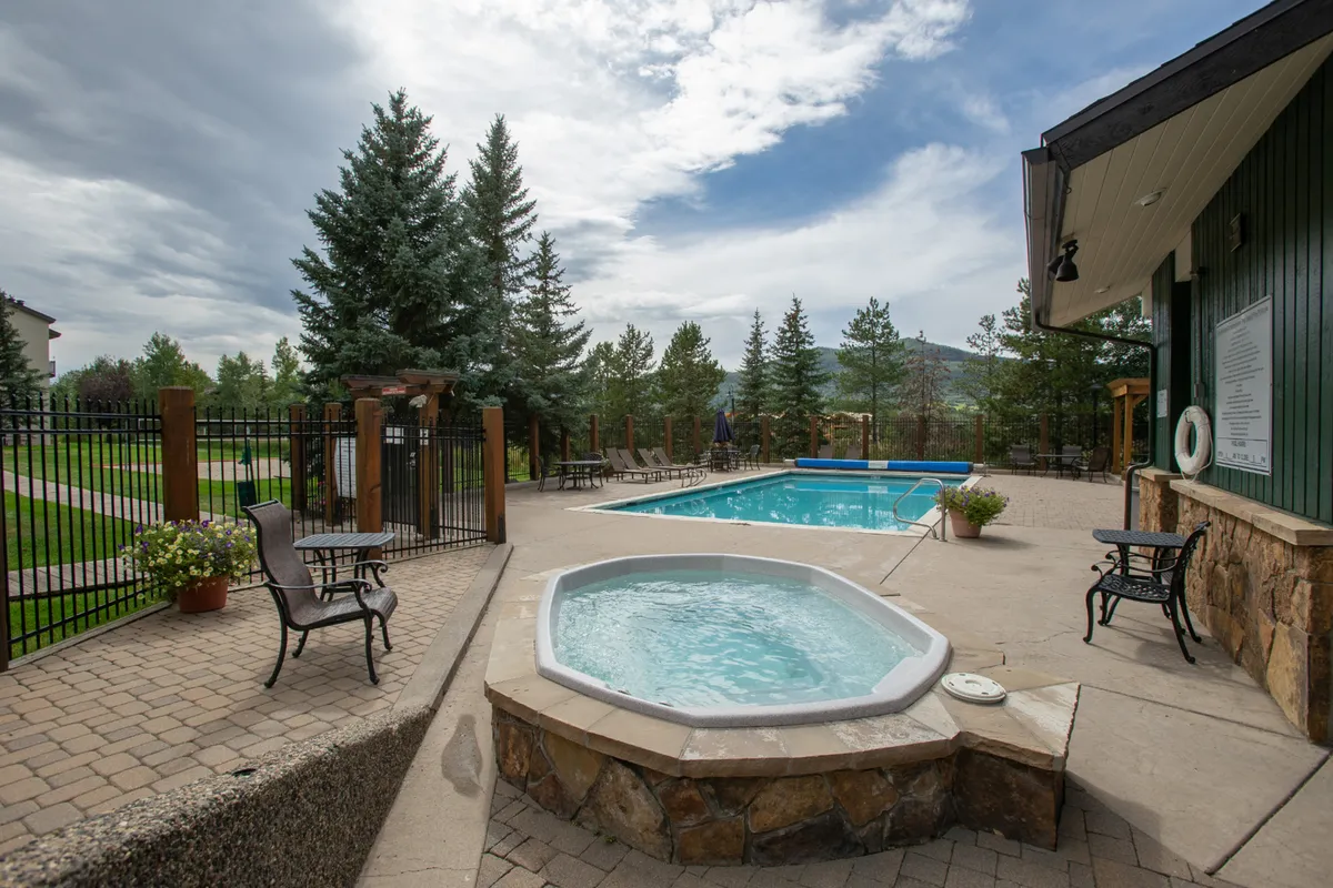 This image displays a sparkling swimming pool accompanied by a hot tub, bordered by mature trees and comfortable lounge chairs. The scene is set outdoors, capturing a tranquil environment suited for both active fun and peaceful relaxation. The area appears perfect for sunbathing, swimming, and unwinding in the hot tub, and the overall setup invites social gatherings.