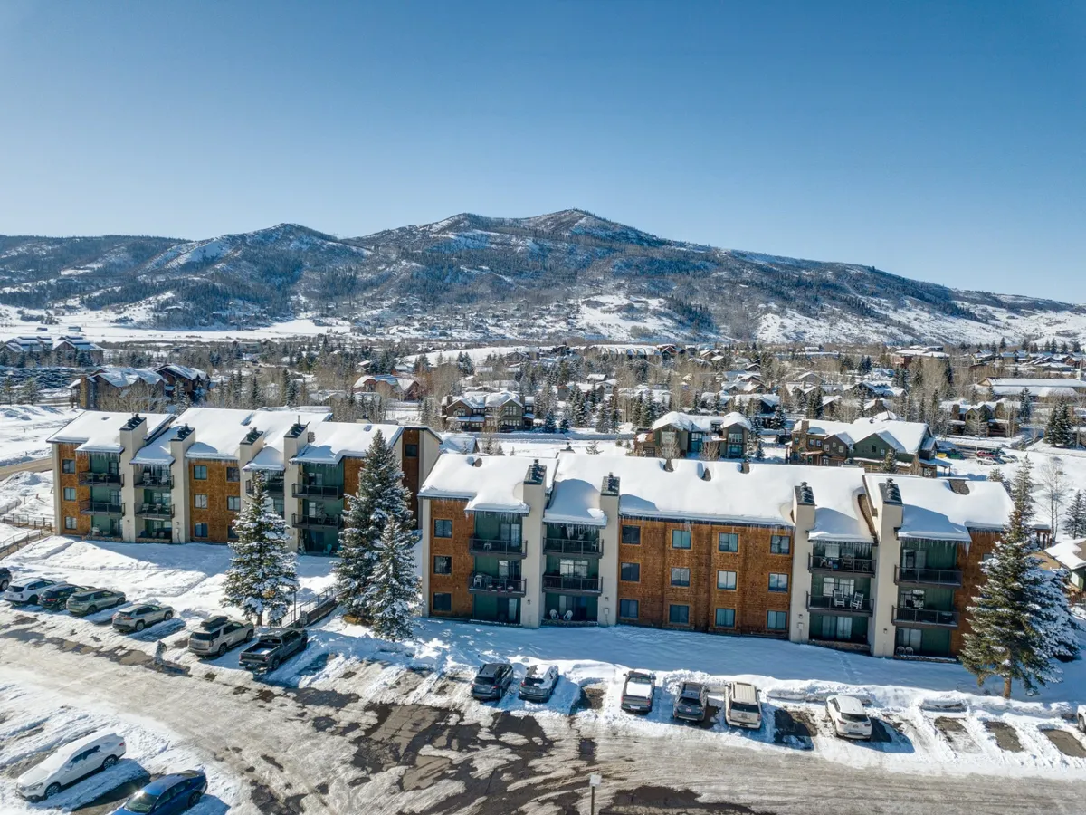 An inviting winter exterior shot with snow-covered buildings, mountain views in the background, and trees framing the property. The scene captures the cozy charm of Steamboat in winter, hinting at nearby slopes and communal winter activities.