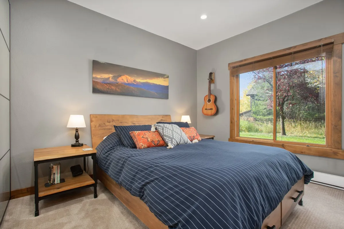 A serene queen bedroom featuring a bed with striped bedding, a large window providing natural light and a scenic view, a guitar on the wall, and tasteful artwork above the bed. The space is styled with modern yet cozy furnishings, inviting relaxation and restful sleep.