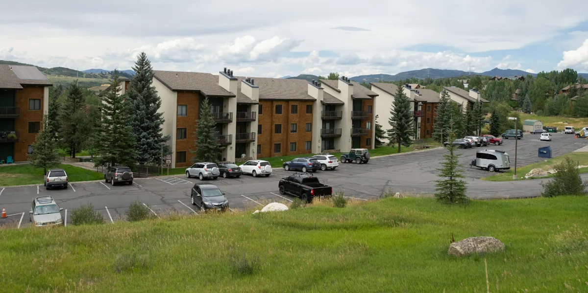 The image features apartment buildings with a backdrop of mountains, green lawns, and a spacious parking area. The area looks ideal for outdoor gatherings, family outings, and easy access to nearby trails.